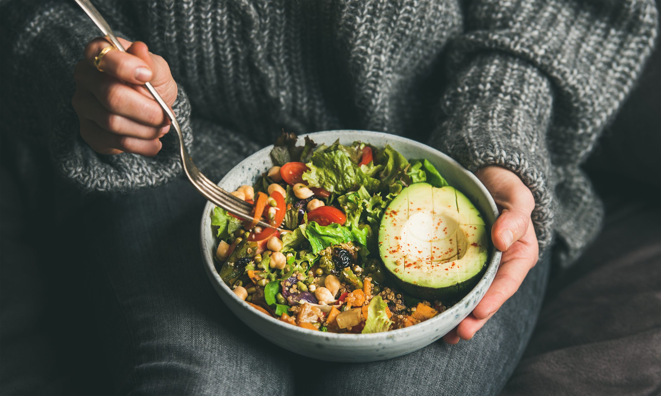 Healthy vegetarian dinner. Woman in jeans and warm sweater holding bowl with fresh salad, avocado, grains, beans, roasted vegetables, close-up. Superfood, clean eating, vegan, dieting food concept