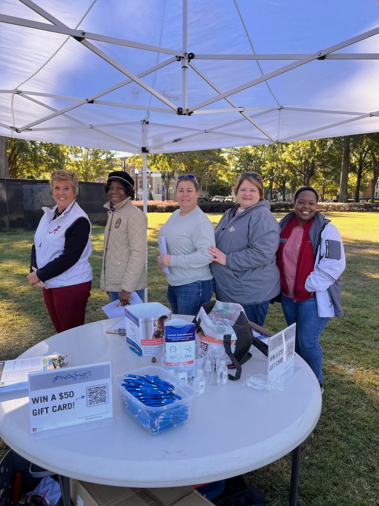 5 women standing side by side smiling behind a table with pens and hand sanitizer on it