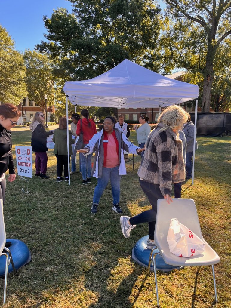 women standing on a bosu ball balancing on one leg holding a chair for balance. There's a white tent in the background and a women directing them