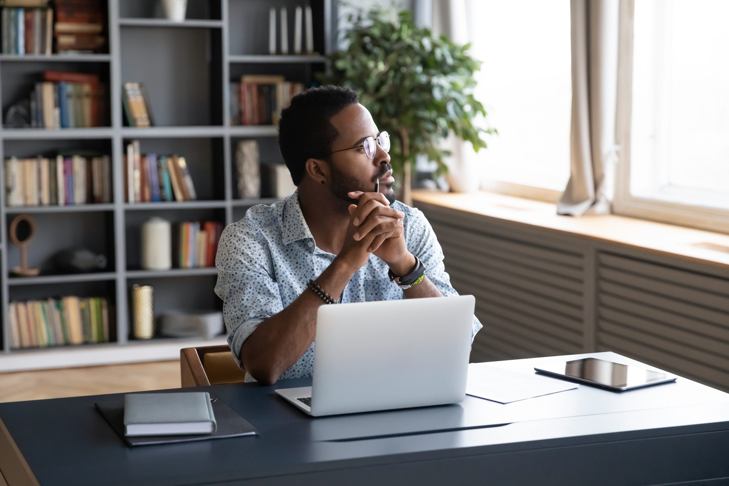 Thoughtful mixed race businessman sitting at table with computer, looking away. Distracted from study job young african american man lost in thoughts, thinking of difficult decision at home office.
