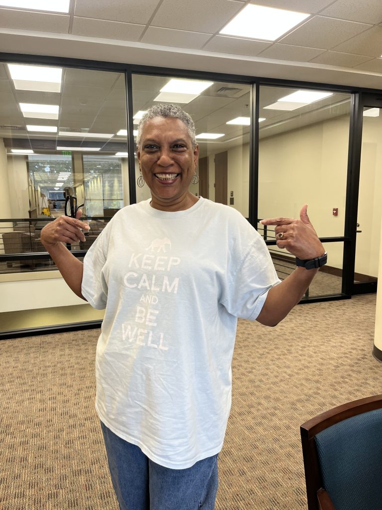 Woman with short gray hair smiling wearing a light blue t-shirt that says "Keep Calm and Be Well"