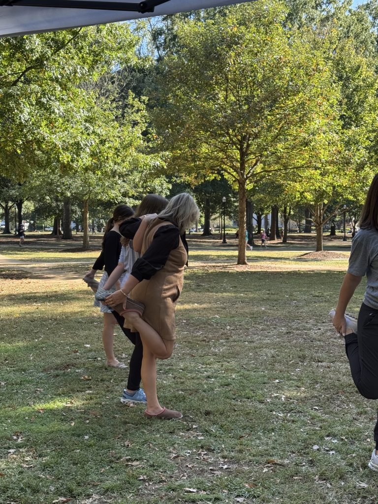 3 women stand in a line facing away from the camera holding one foot behind them to stretch their quads. They hold onto each other for balance