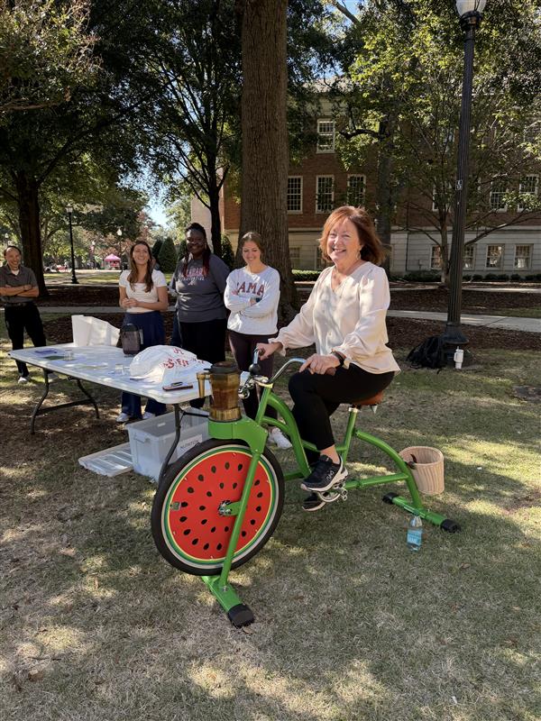 A women sits on a stationary bike painted green with the front wheel looking like a watermelon while other students look on a smile