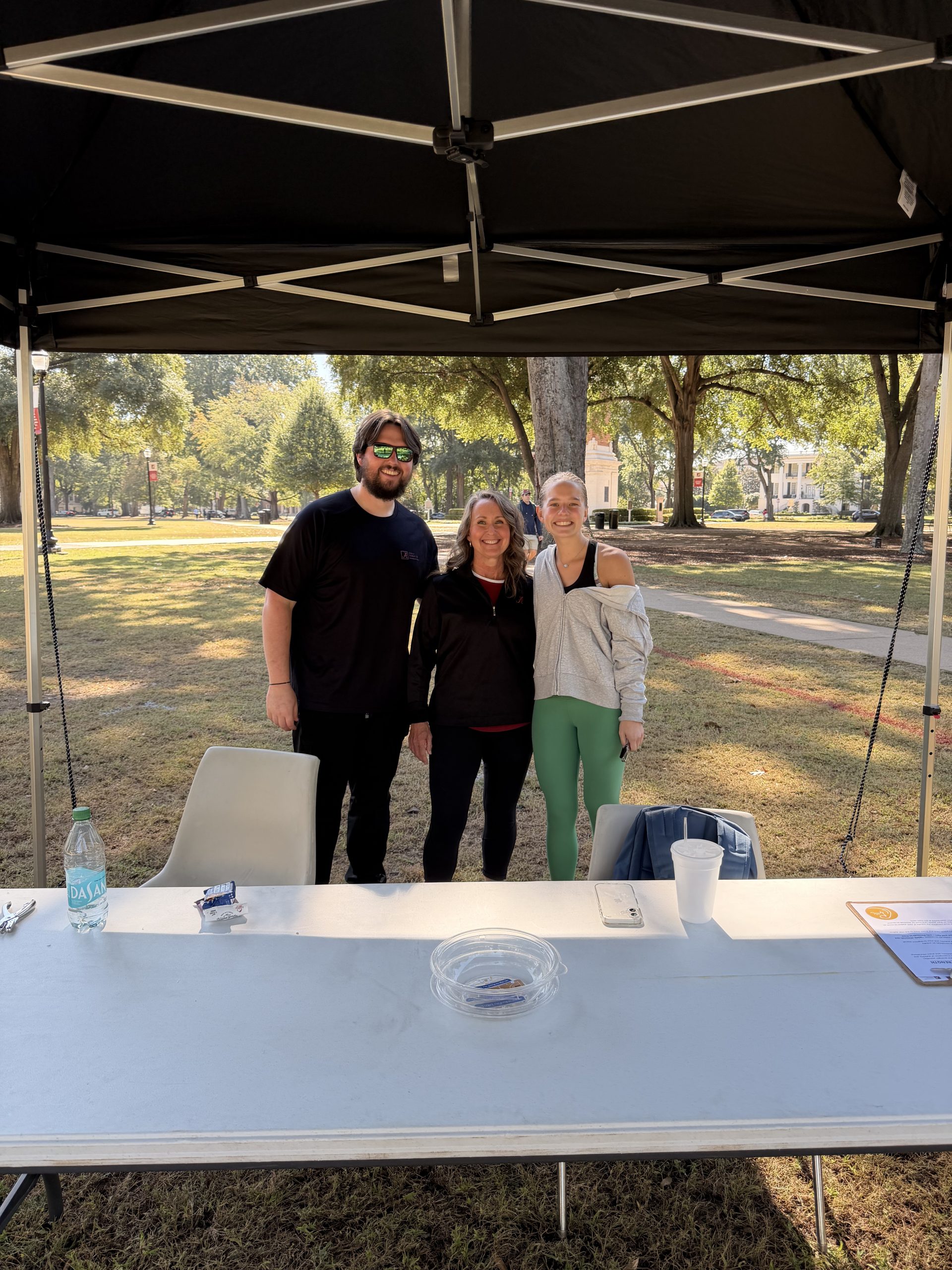 a man and two women stand behind a table outside under a tent smiling