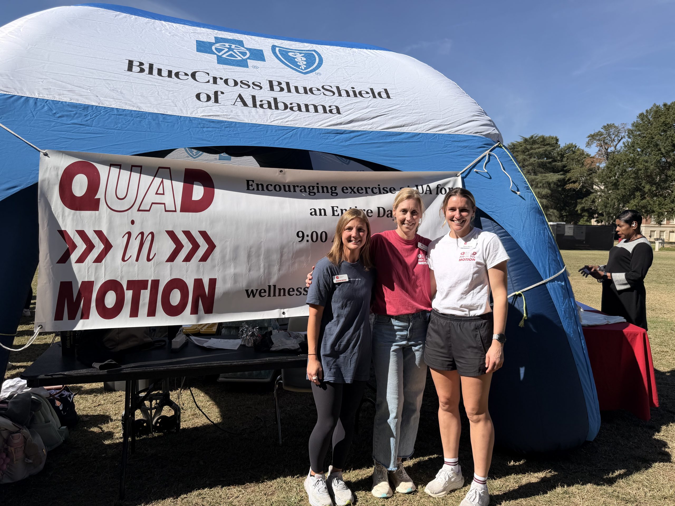 The wellness and worklife team stand in front of a banner that says Quad in Motion outside under an inflatable tent