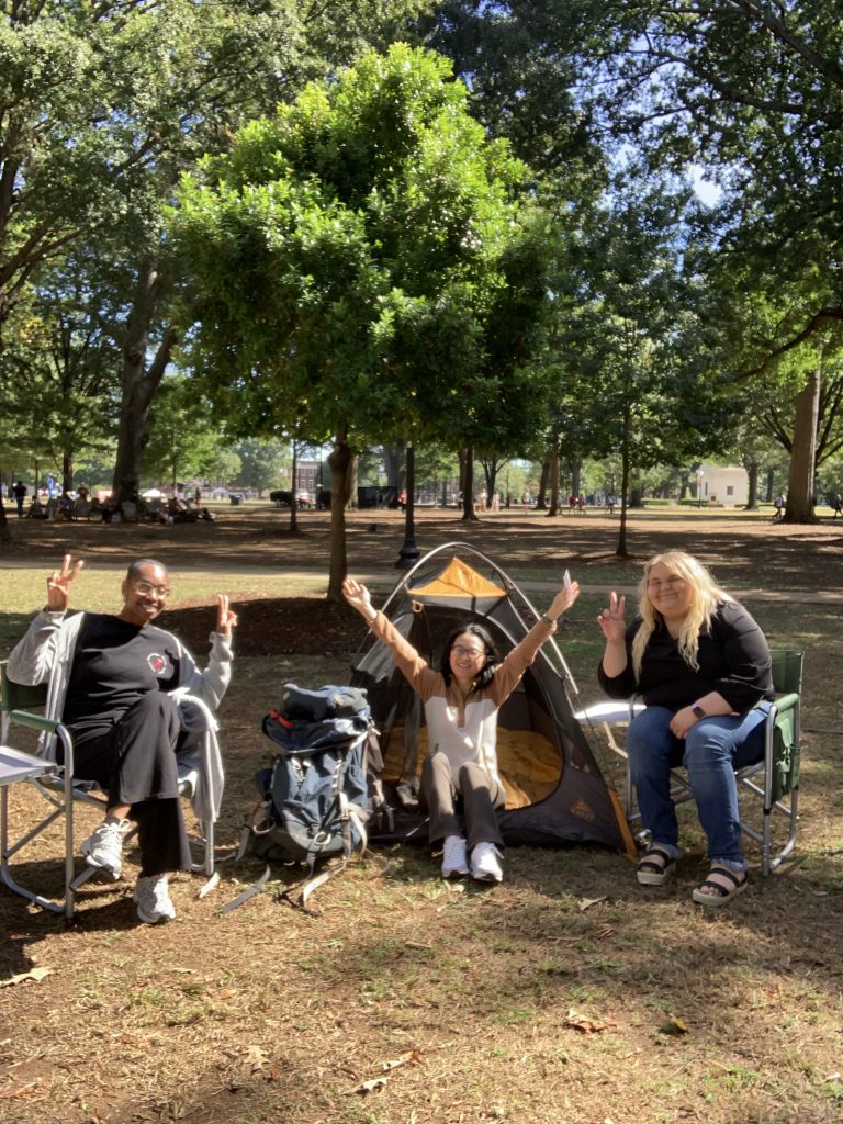 3 women sit in a camping set up outdoors holding their arms overhead and peace signs